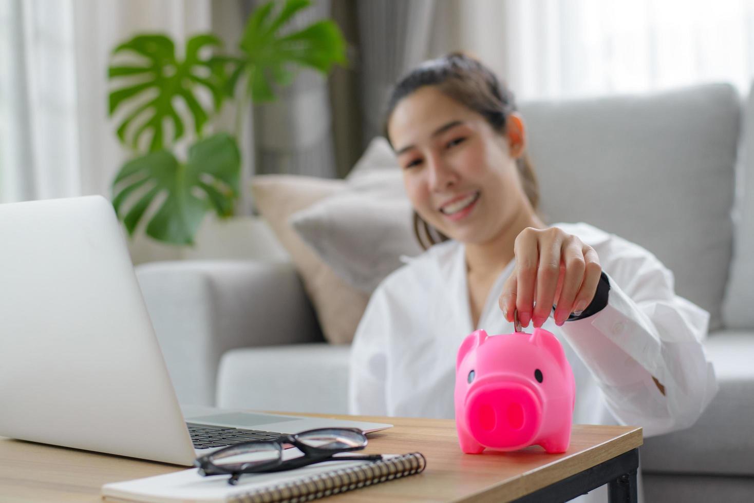 mujer sonriente con camisa blanca sentada frente al sofá en la sala de estar usando la mano poniendo dinero en la alcancía para ahorrar dinero para la inversión y la jubilación. riqueza y concepto financiero. foto