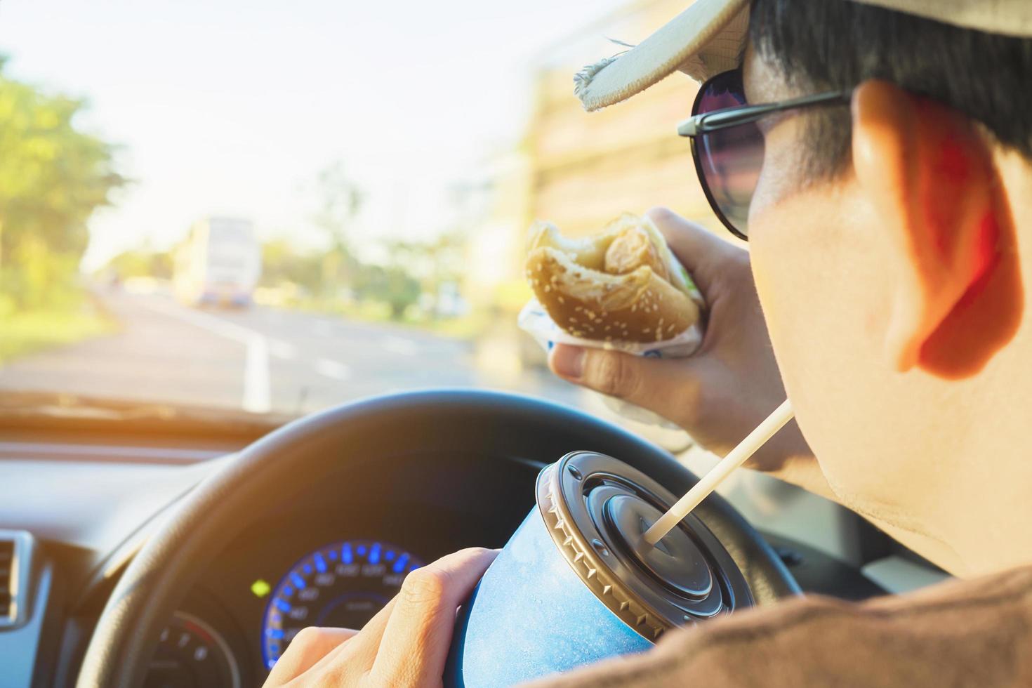 el hombre está comiendo peligrosamente un perro caliente y una bebida fría mientras conduce un automóvil foto