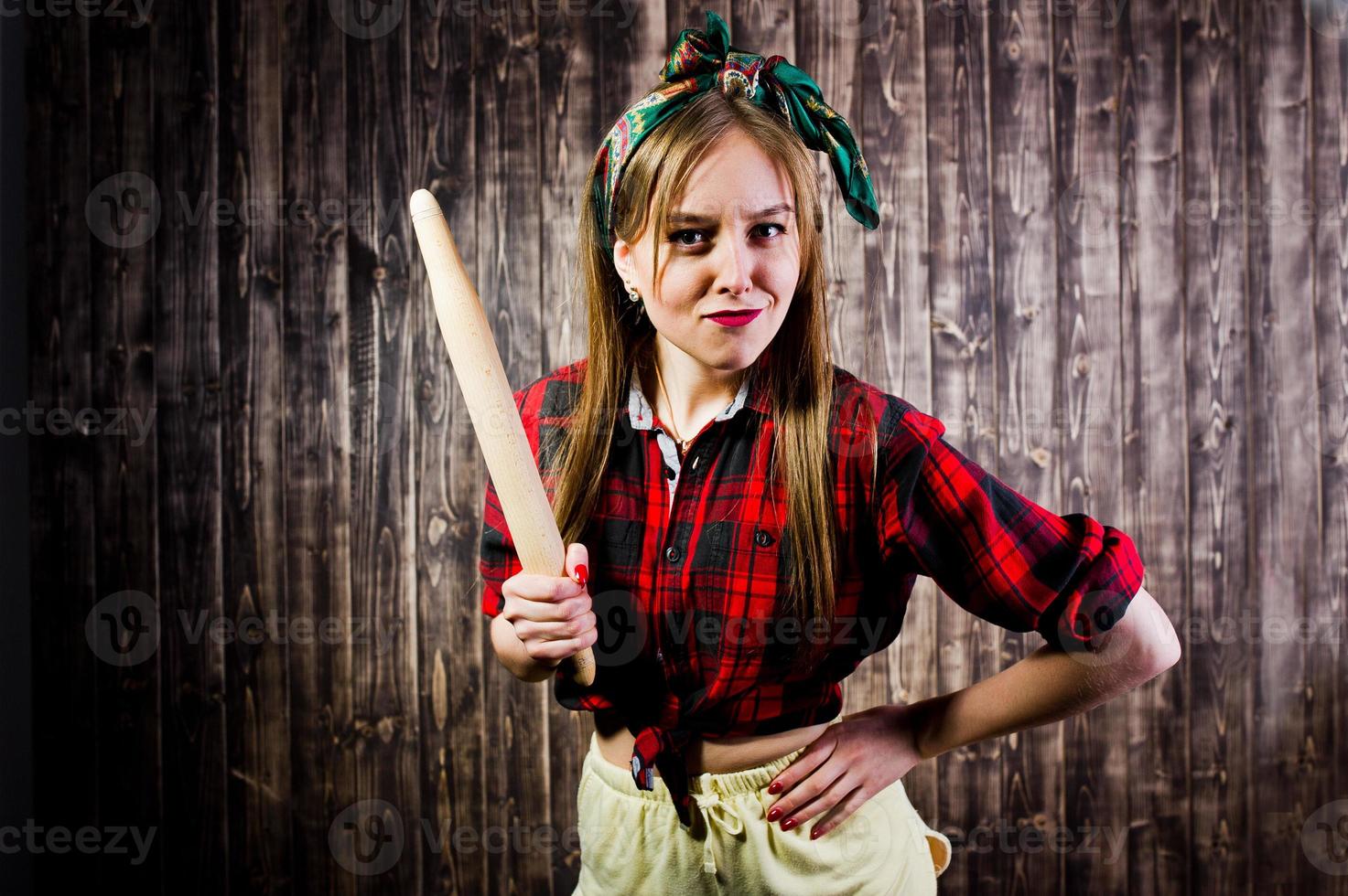 Young funny housewife in checkered shirt and yellow shorts pin up style with kitchen rolling pin on wooden background. photo