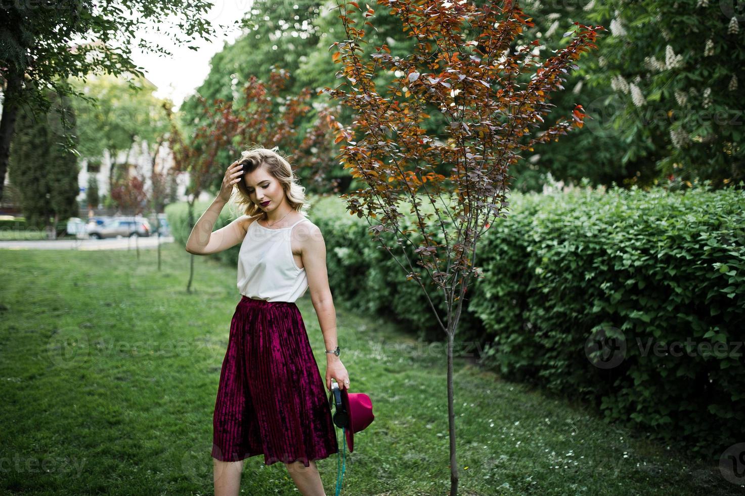 Fashionable and beautiful blonde model girl in stylish red velvet velour skirt, white blouse and hat, posed at park with phone and earphones. photo