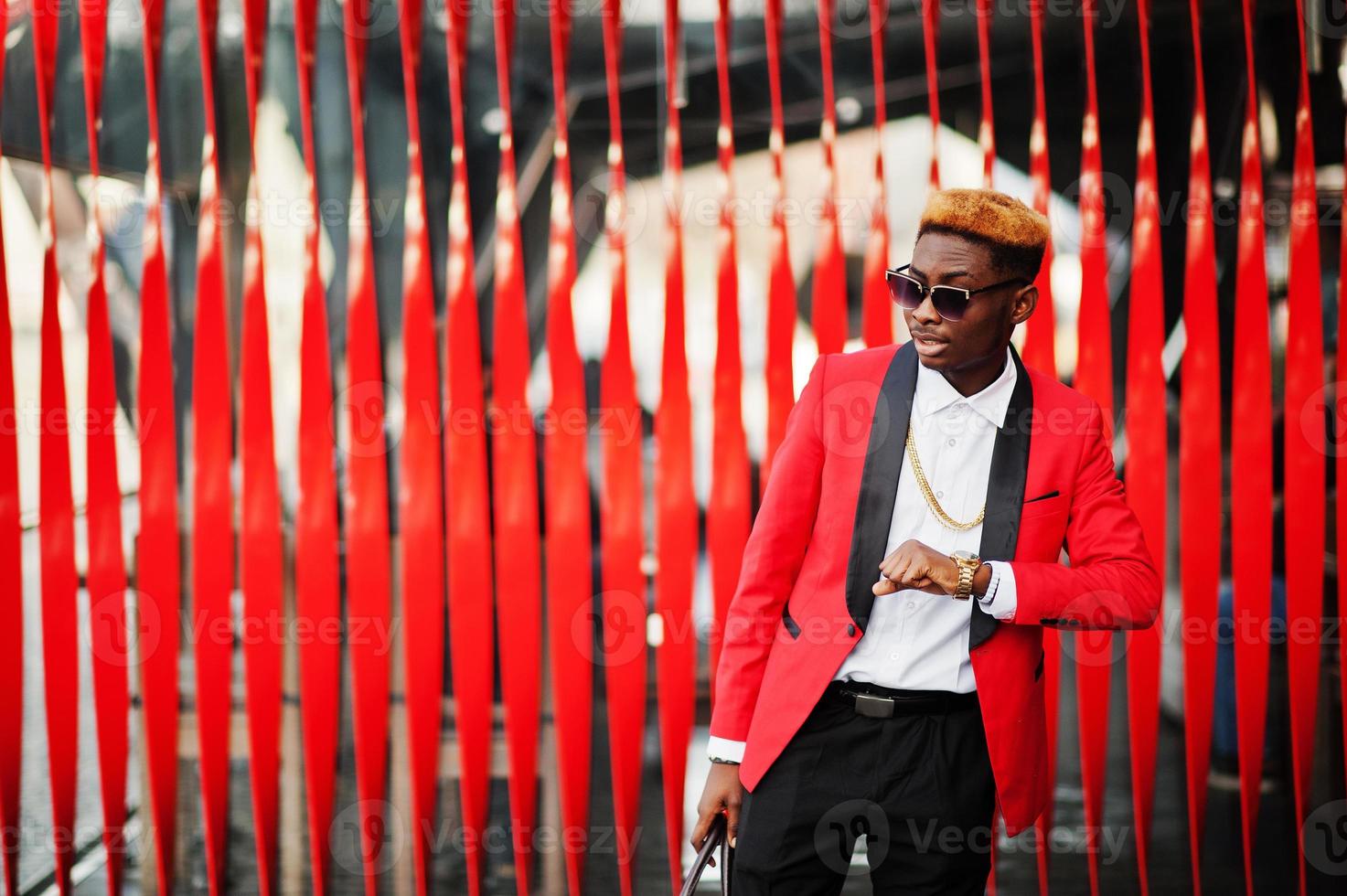 Fashion african american man model at red suit, with highlights hair and handbag posed at street and looking at his watches. photo