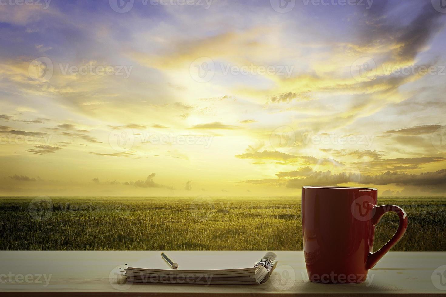 a red cup of coffee and note pad on wooden table and Empty grassland and sky at evening time photo