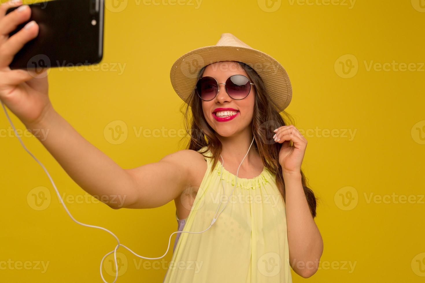 Close up portrait of happy brunette woman in yellow blouse and hat making selfie on yellow background. photo