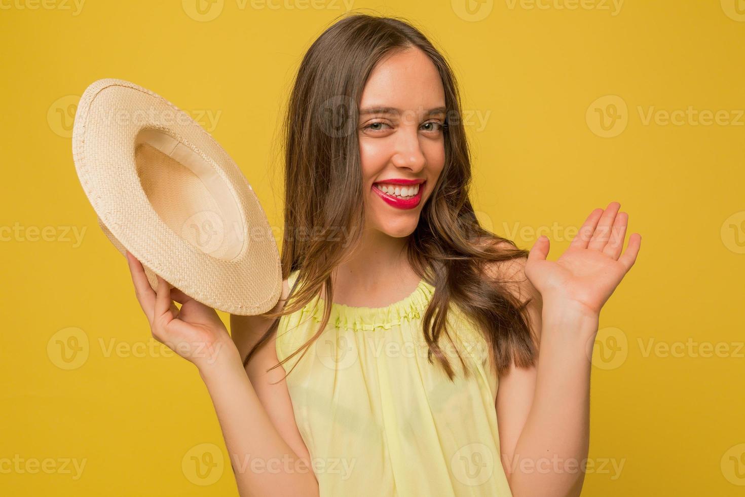 Funny charming girl with pink lips is waiving at camera while holding hat and posing at camera over yellow background photo