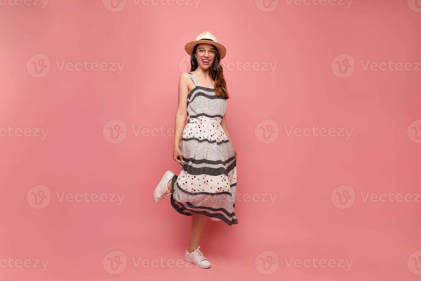 espectacular modelo femenino elegante con peinado largo saltando con cara feliz sobre fondo rosa. adorable chica con sombrero de verano y vestido brillante expresando emociones positivas. foto