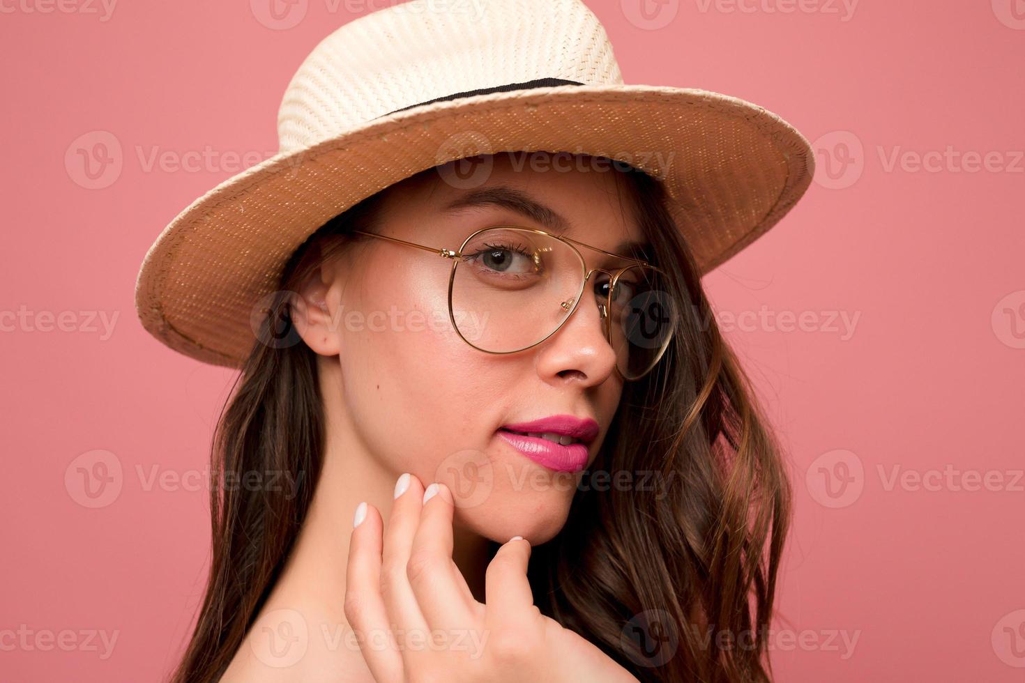 Close up tender portrait of lovable girl with great make up wearing glasses and hat is looking at camera over pink background photo