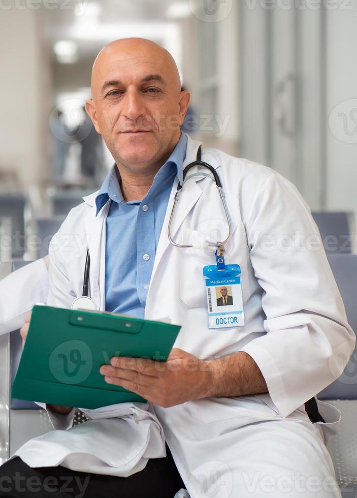 Male medical doctor person sits in clinic, portrait. Professional specialist man proudly working in hospital. Mature practitioner wears white uniform and stethoscope confident in healthcare occupation photo