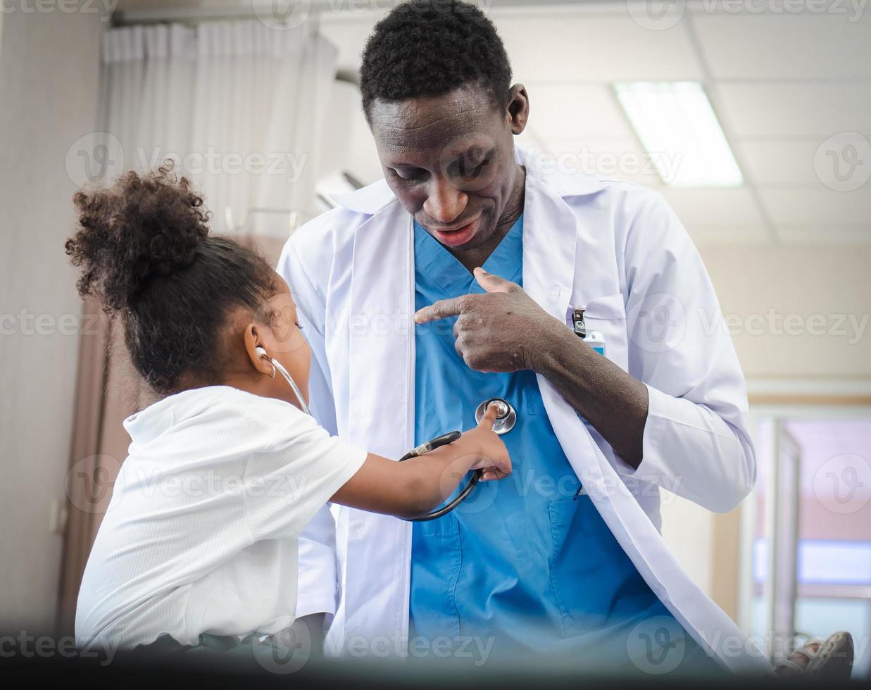 Selective focus at doctor person playing with cute afro child patient in hospital ward. Friendly pediatrician entertaining girl kid enjoying with stethoscope for heartbeat during medical exam. photo