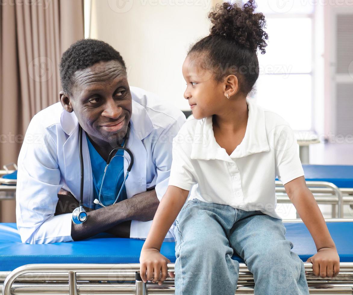 Child patient sitting on hospital bed with African doctor for medical care. Smiling little girl happy healthy after professional checkup at clinic. Practitioner, pediatrician checking, examining kid photo