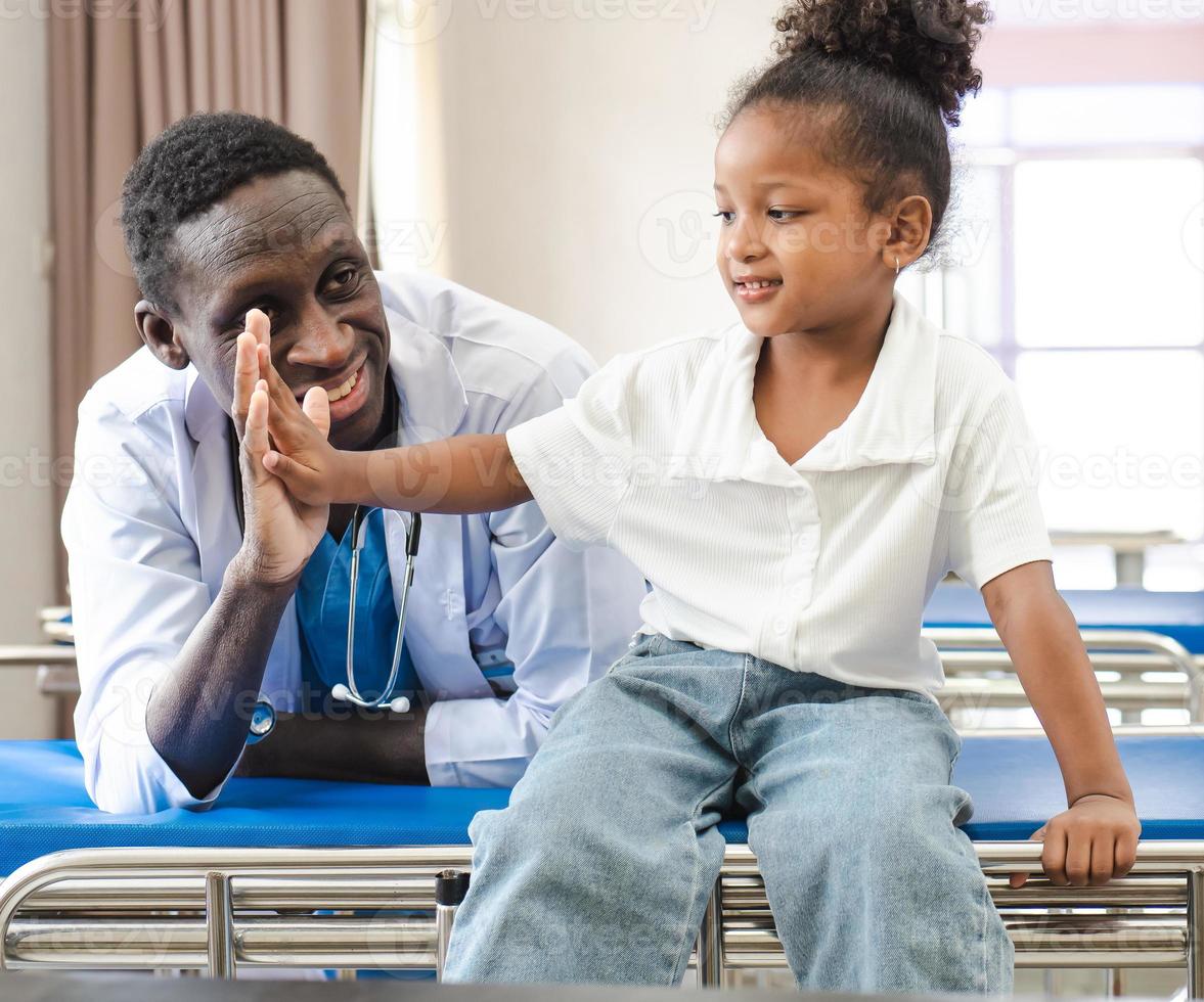 Child patient sitting on hospital bed with African doctor for medical care. Smiling little girl happy healthy after professional checkup at clinic. Practitioner, pediatrician checking, examining kid photo