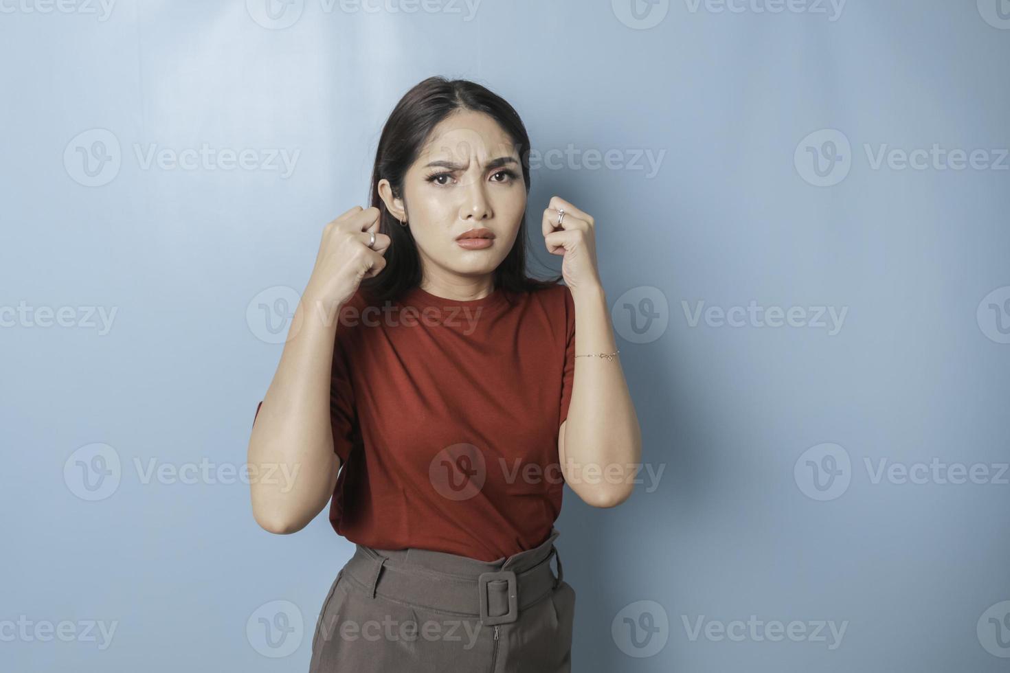 A dissatisfied young Asian woman looks at the camera posing on a blue background, disgruntled girl wearing red t-shirt irritated face expressions show negative attitude concept image photo