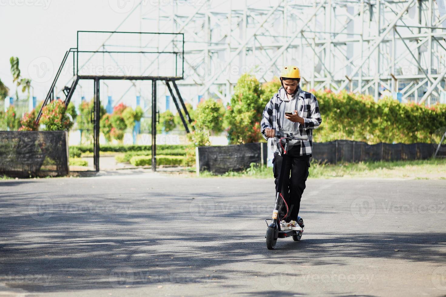 Asian man riding electric scooter while looking at the phone on park street. Modern and ecological transportation concept. photo
