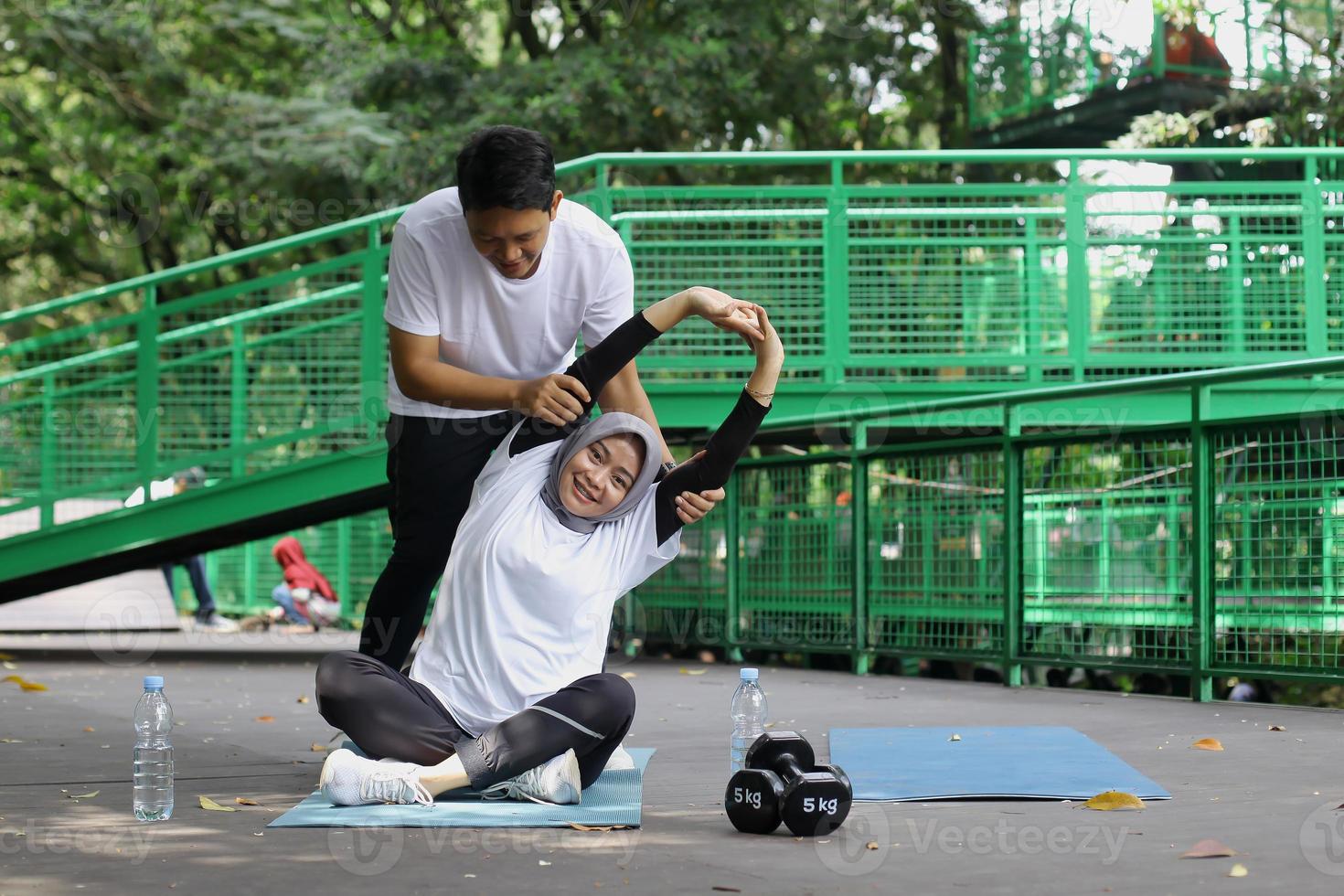 Young Asian muslim couple doing yoga practice together. Man helping woman to do a yoga pose at park. photo