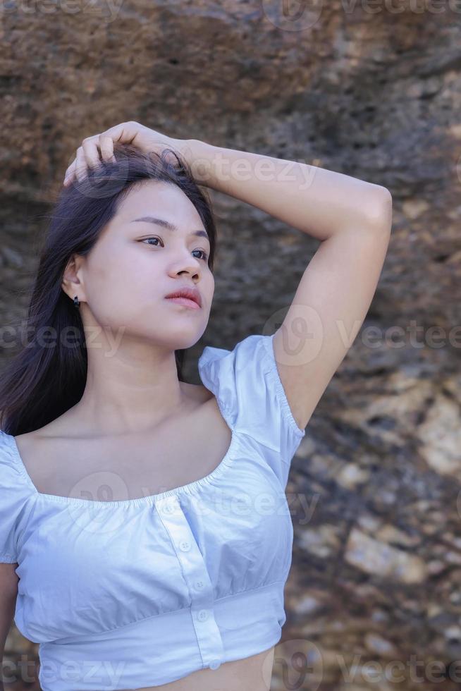 retrato de hermosa mujer de moda en bikini blanco posando en la playa de roca. joven asiática vestida con traje de baño sobre una roca en un día de verano con fondos de puesta de sol en koh larn en pattaya foto