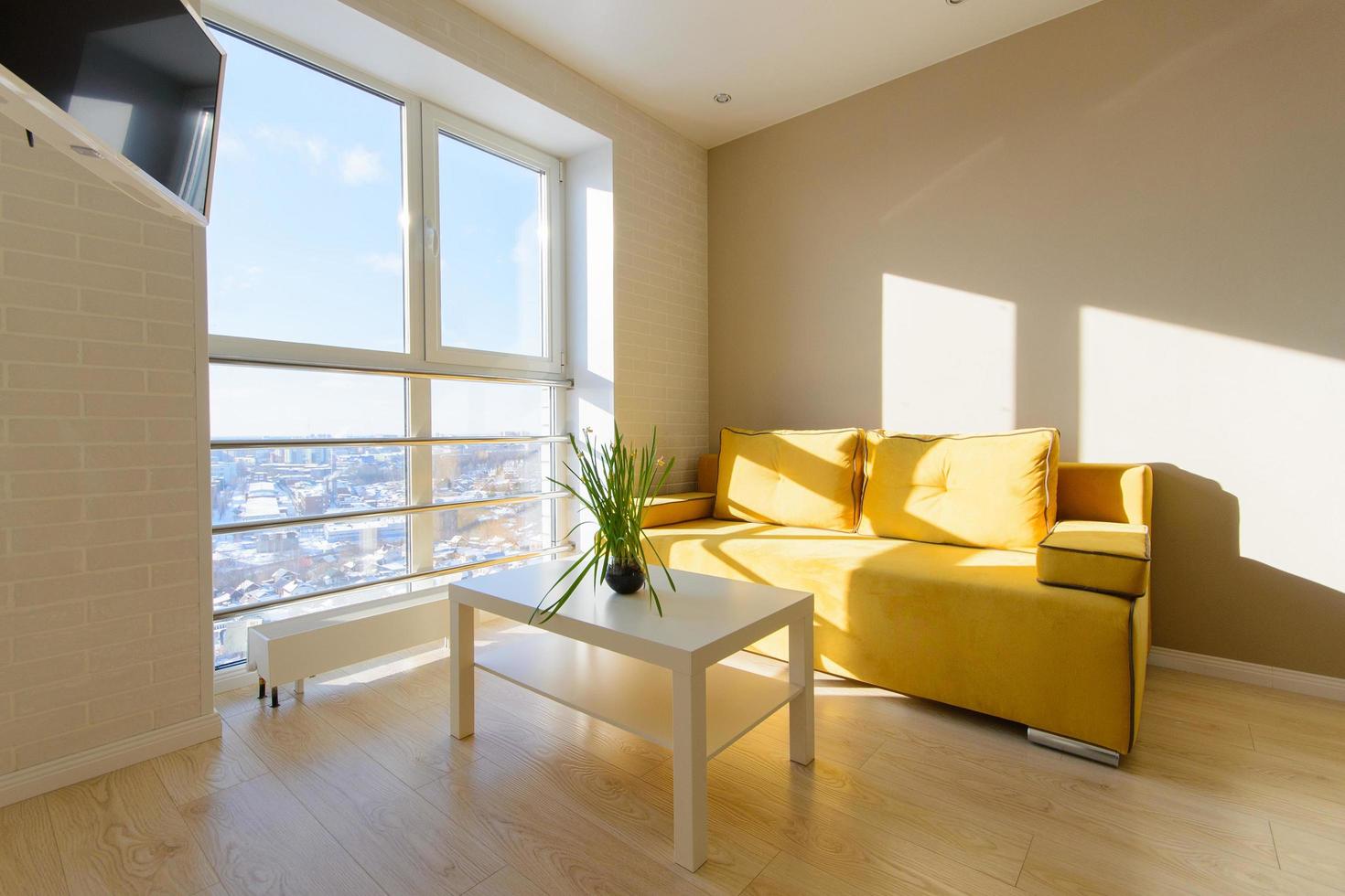 Modern cozy apartment interior, living room with yellow sofa, white coffee table and tv on wall, panoramic window with beautiful view to the city. photo