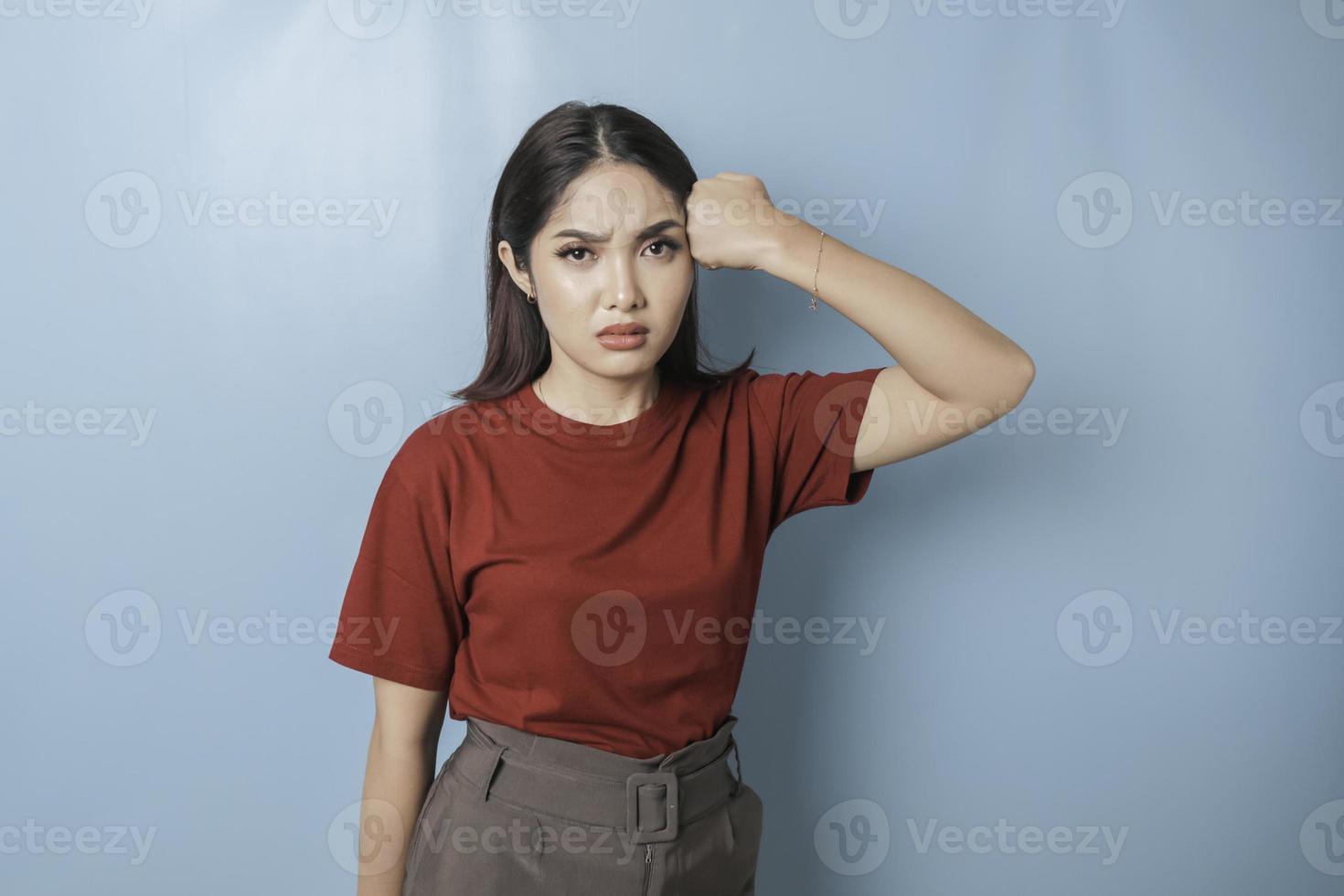 A dissatisfied young Asian woman looks at the camera posing on a blue background, disgruntled girl wearing red t-shirt irritated face expressions show negative attitude concept image photo
