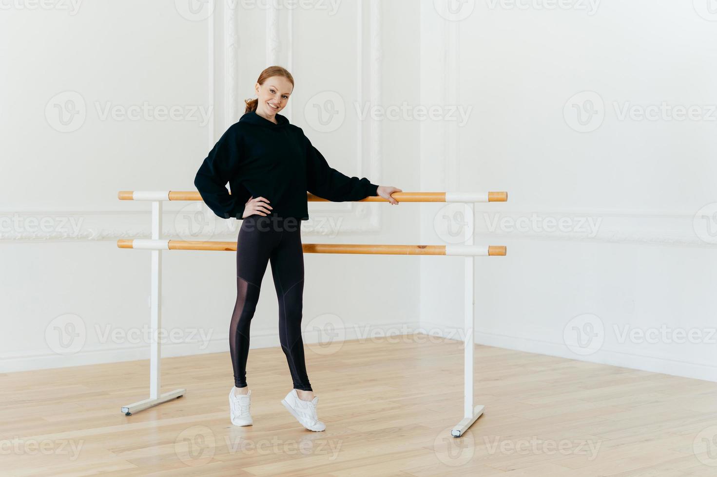 Full length shot of cheerful redhead slim female stands on tip toes near ballet barre, has pilates exercises, keeps fit, holds hand on waist, wears black sport clothes, poses in spacious white hall photo