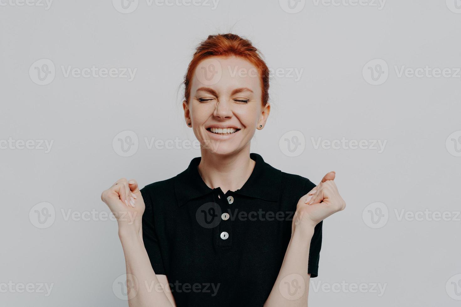 Happy euphoric red haired female with closed eyes holding clenched fists up and celebrating win photo