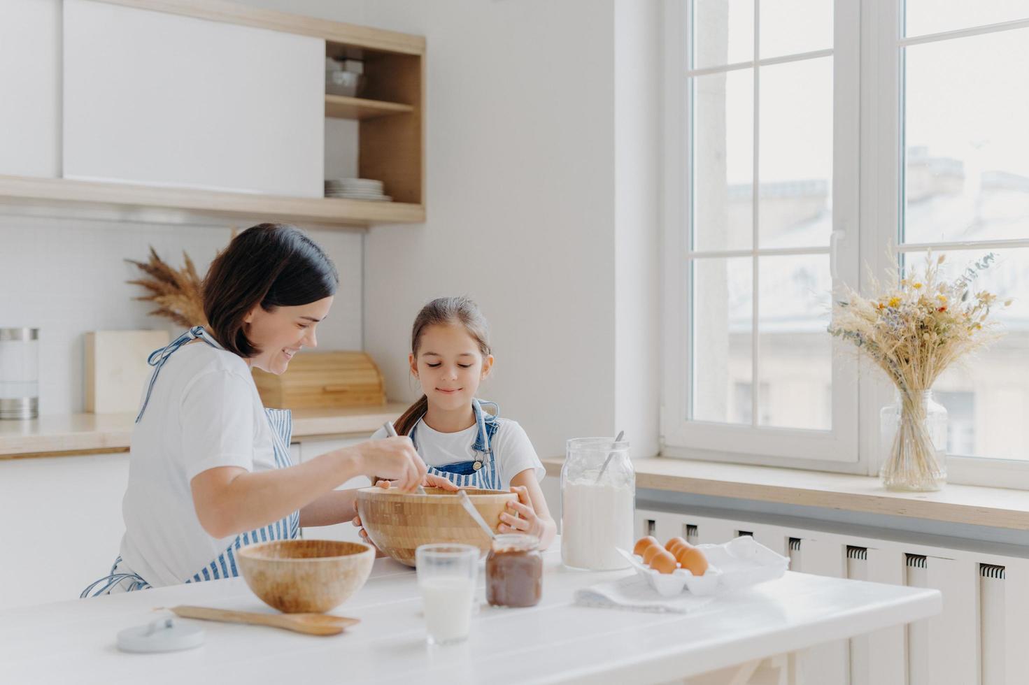 Brunette woman with smile shows little daughter how to cook, gives culinary lesson, bought different products in shop for cooking, wear aprons, smile pleasantly, window with vase, kitchen interior photo