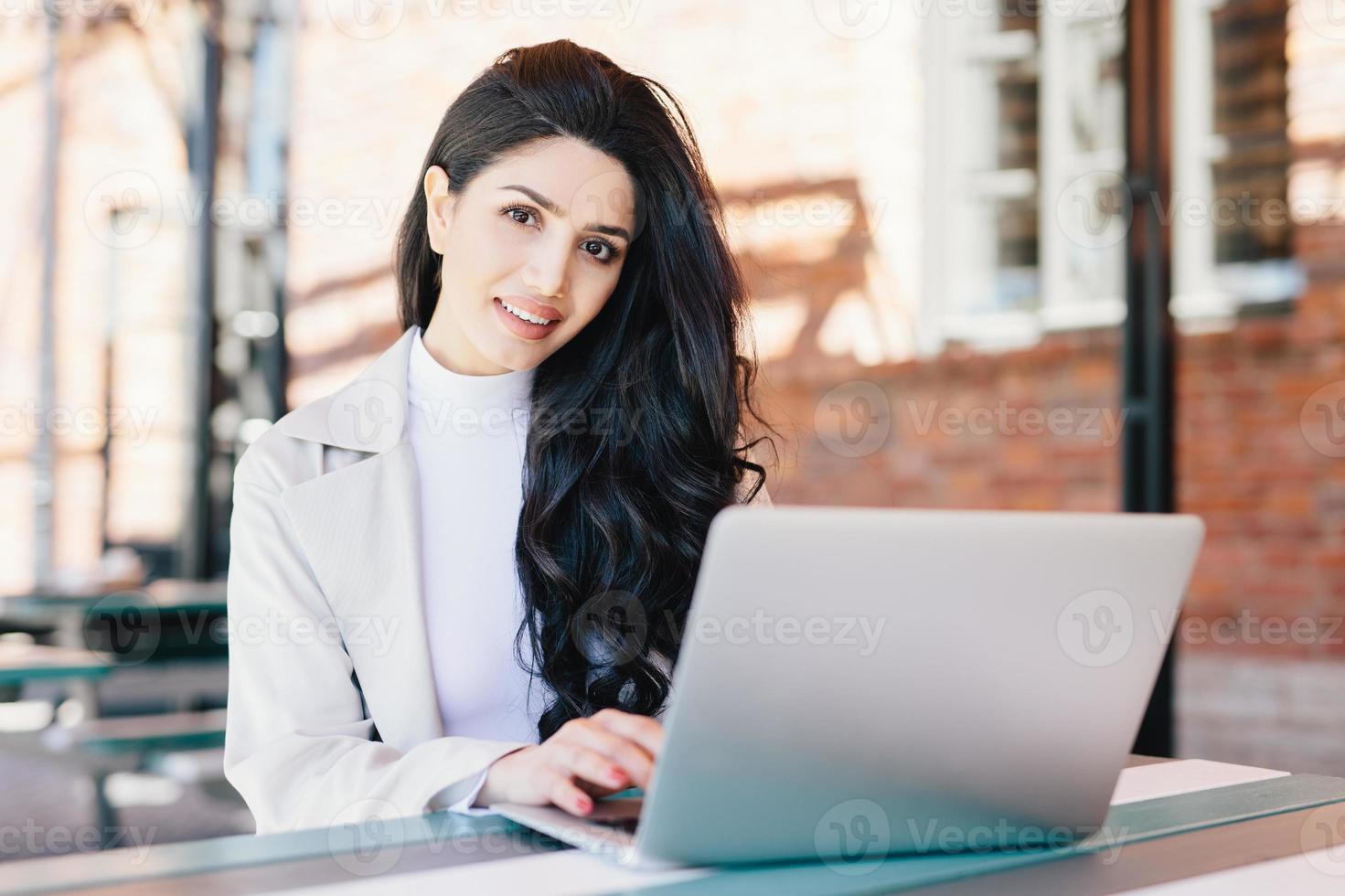 concepto de tecnología y comunicación. exitosa mujer de negocios europea con hermosa apariencia trabajando en un café en una computadora portátil con expresión sonriente mirando en cámara mientras se sienta al aire libre foto