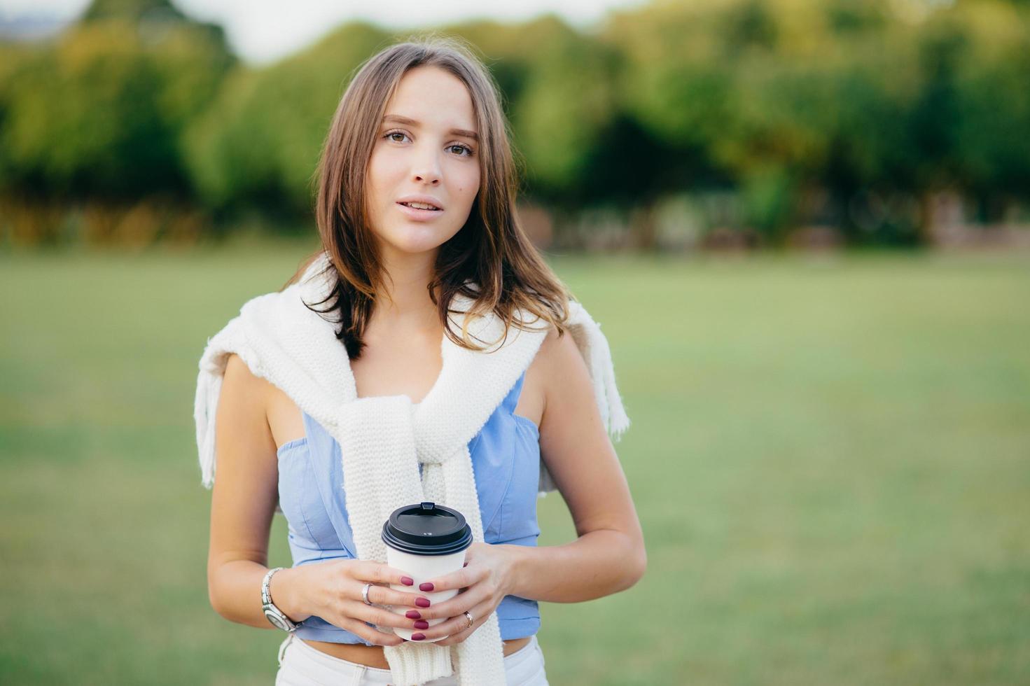 Pretty female model has dark hair, dressed in casual clothes, has manicure, holds takeaway coffee, enjoys sunny warm weather, beautiful nature, recreation time, talks with friends have picnic together photo