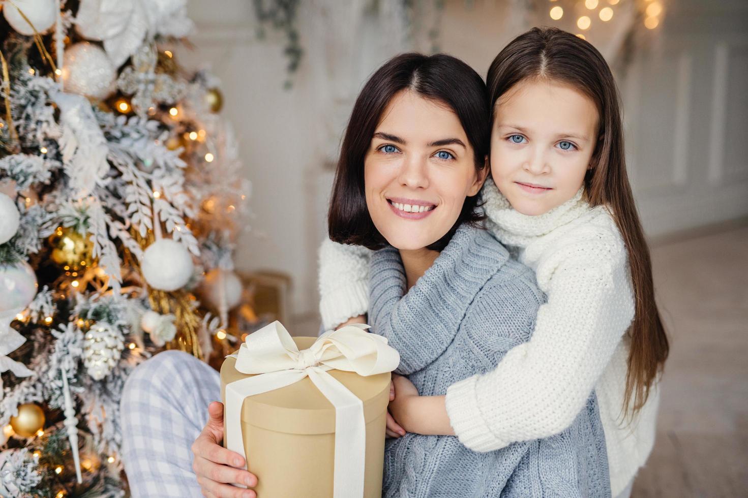 Adorable blue eyed small kid huggs with great love her mother who holds wrapped gift box, stand near decorated New Year tree, happy to celebrate winter holidays. People, celebration, presents concept photo