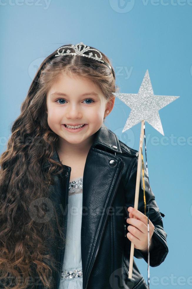 Satisfied small female child has long curly hair, dressed in black leather jacket, wears crown, holds magic wand, isolated over blue background. Dark haired positive kid anticipates for miracle photo