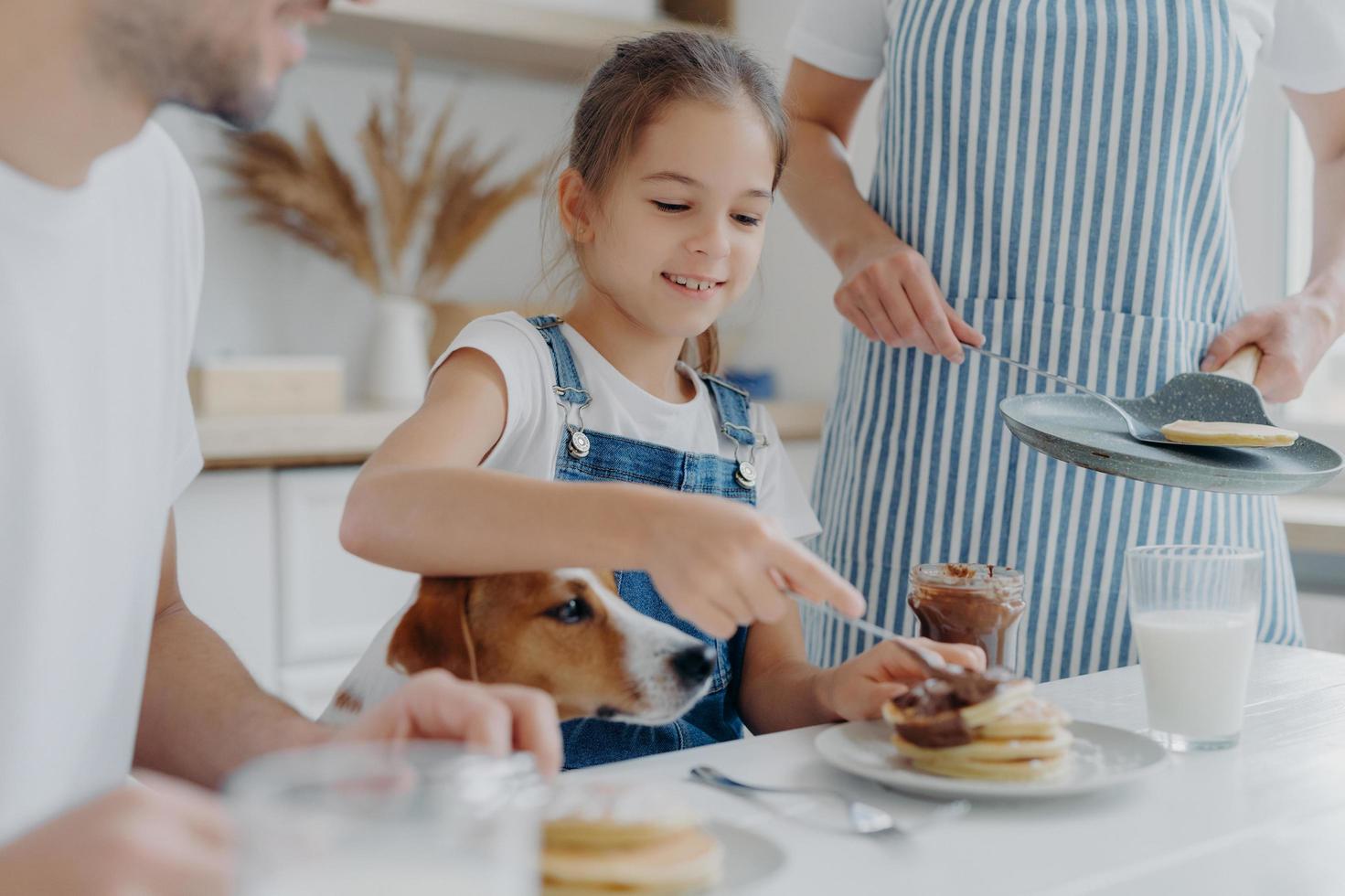 Small kid, her father and dog sit together at kitchen table, eat freshly prepared pancakes, mother in apron stands near holds pan. Family appetizing tasty desset at kitchen. Cooking, nutrition concept photo