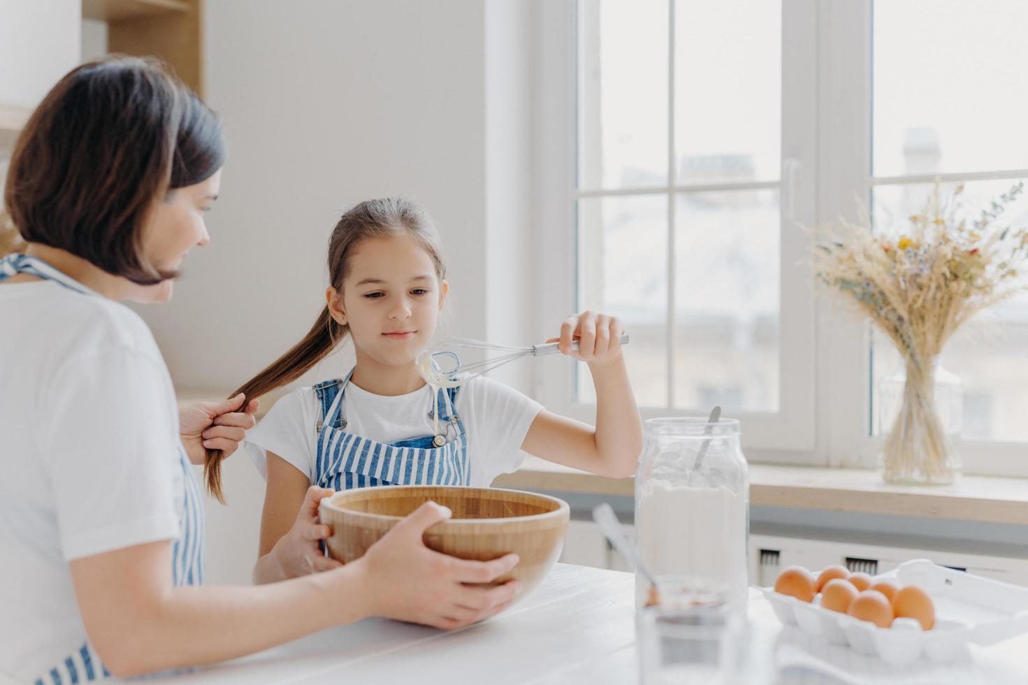 Shot of small kid helper whisks cream with beater, learns how to bake, listens useful advice from mother who holds her pony tail, sit at white kitchen table together. Family cooking together photo