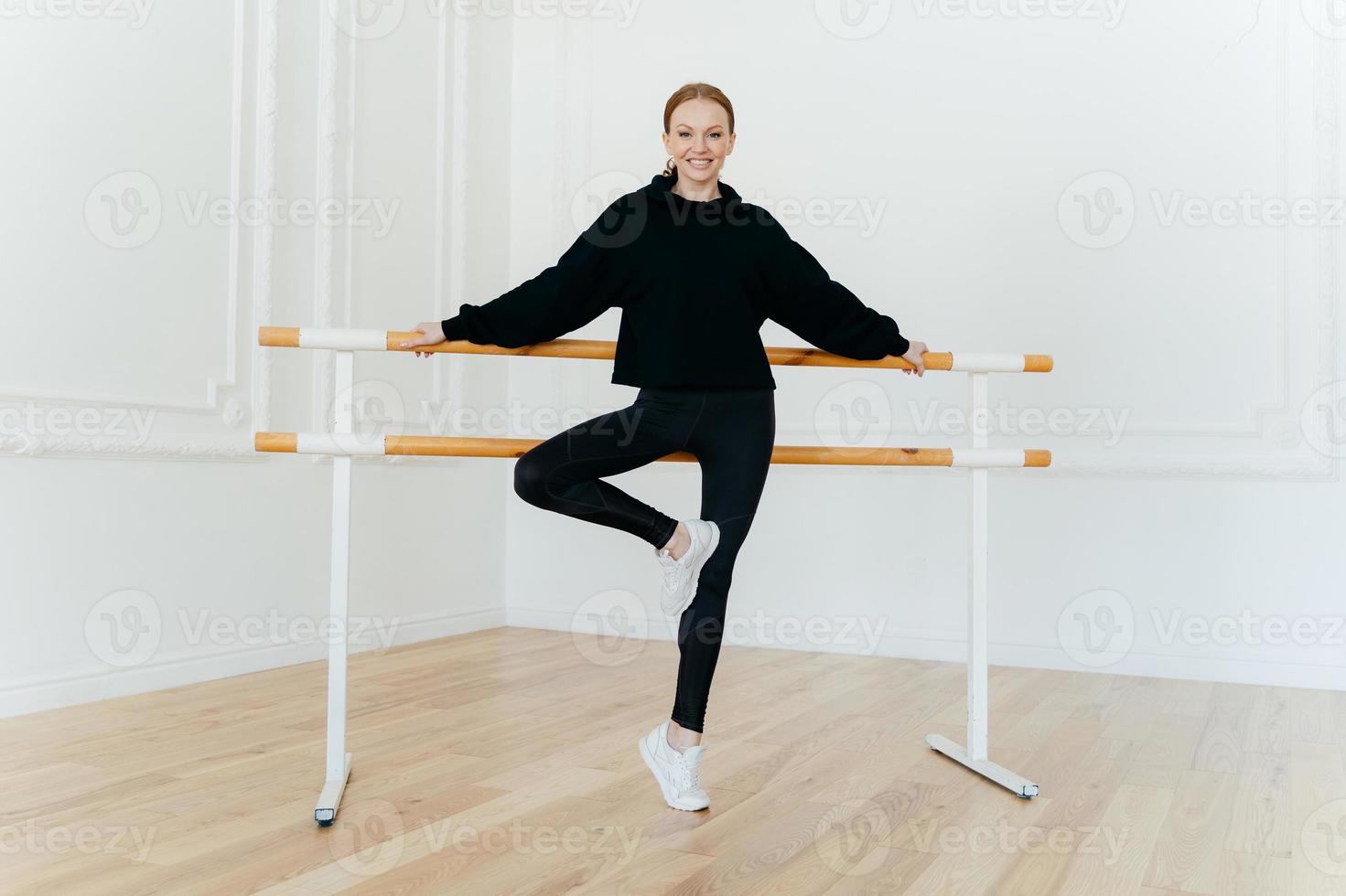 Horizontal shot of flexible young female leans on ballet barre, wears black costume and white sportshoes, smiles positively, poses in dancing hall. Ballet instructor demonstrates different exercises photo