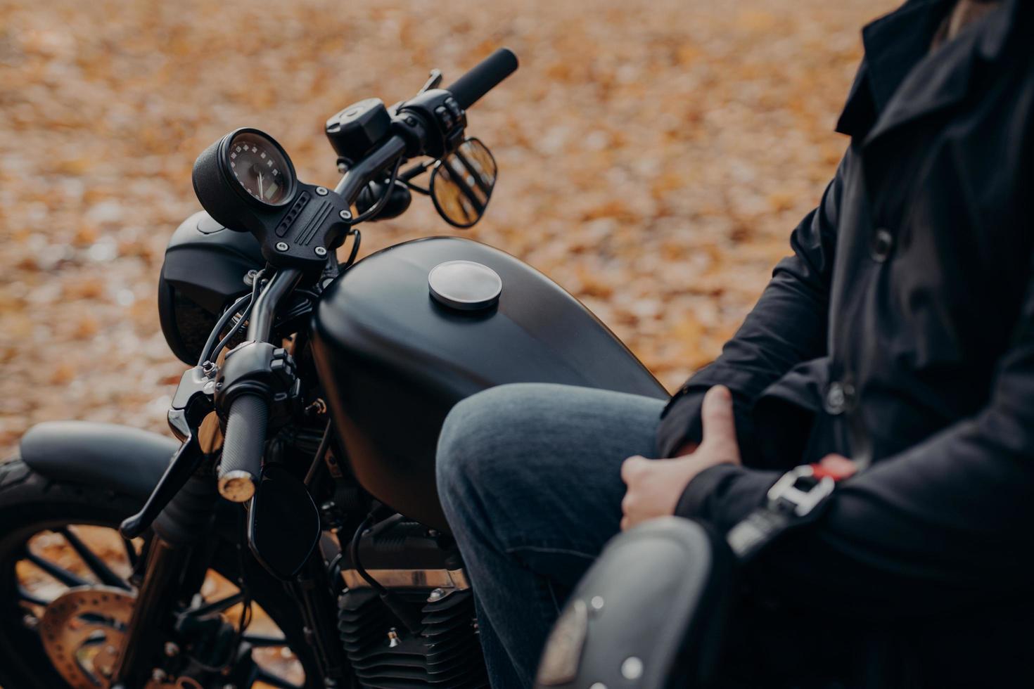 Cropped shot of faceless biker poses on fast motorbike, stops in park, ground covered with fallen leaves, enjoys travel and journey. People, lifestyle, transport concept. photo