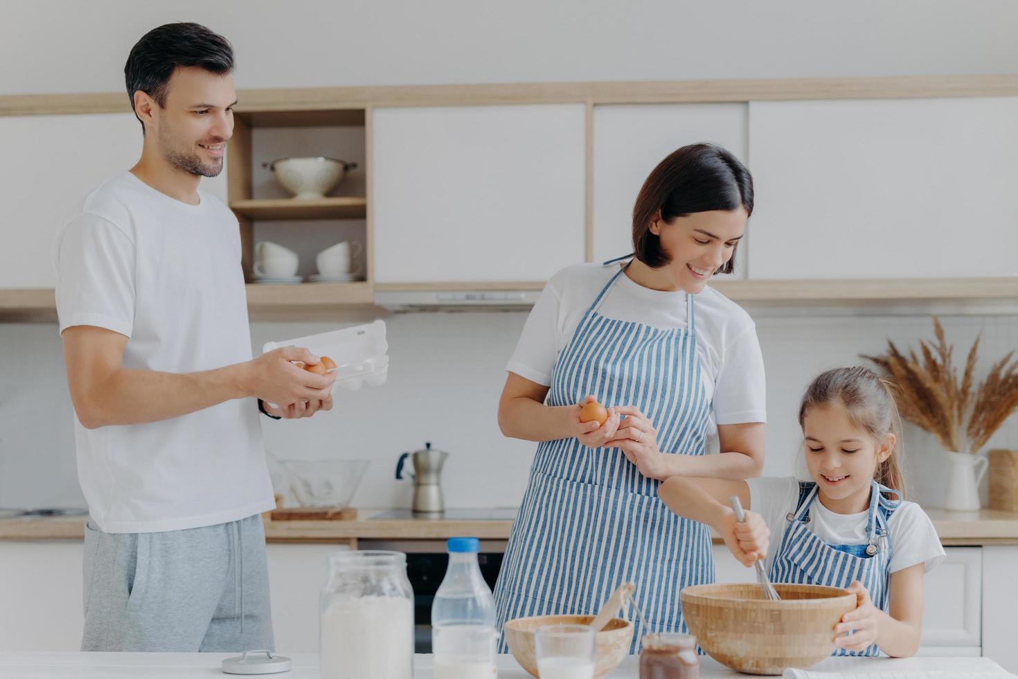 los padres felices cocinan juntos con su hija, posan en la cocina casera moderna dan huevos para agregar a la masa, los niños pequeños baten los ingredientes en el tazón tienen expresiones alegres. concepto de comida y familia. cocinando el desayuno foto