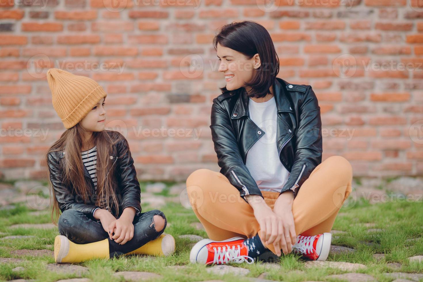 Shot of pleased realxed two sisters pose crossed legs, sit on lawn, pose against brick wall background, have happy facial expressions. Little kid in leather jacket spends time with her mother photo