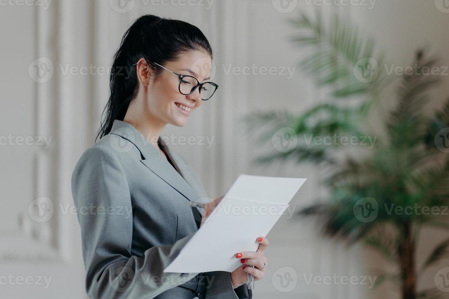 Glad female lawyer writes in corporate paper documents, wears optical glasses, formal clothes, stands in own cabinet, develops new strategy, has happy expression. People, career, employment concept photo