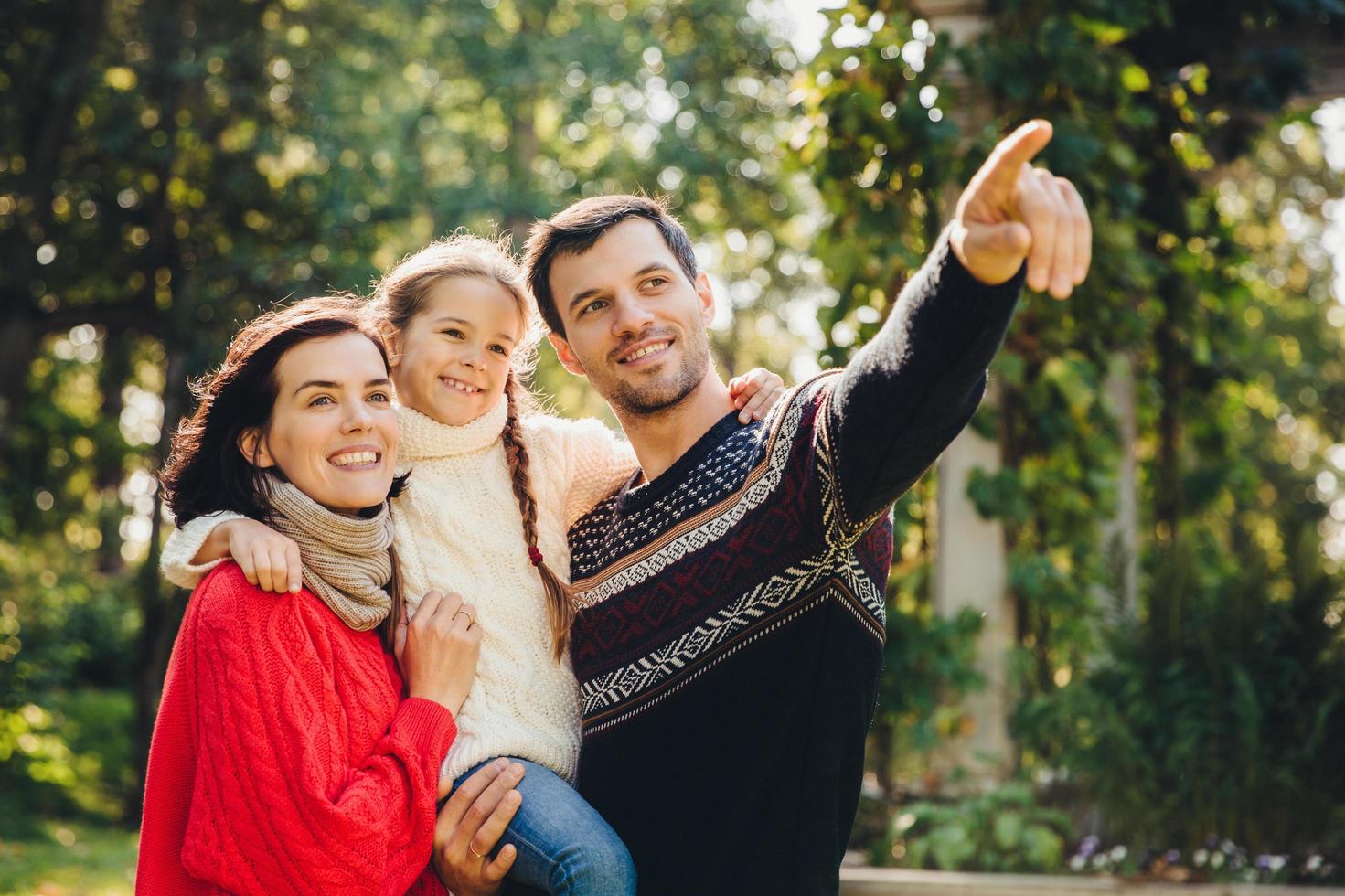 retrato al aire libre de una familia feliz y sonriente que camina juntos. el padre afectuoso le muestra algo a su pequeña hija en la distancia. la familia admira el amanecer, la hermosa naturaleza, usa ropa abrigada foto