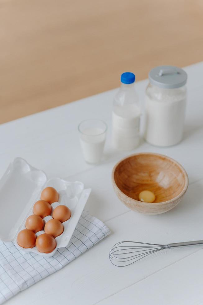 Top view of different dough recipe ingredients at kitchen table. Eggs, whisk, glass of milk and flour. Cooking, baking and culinary concept. photo