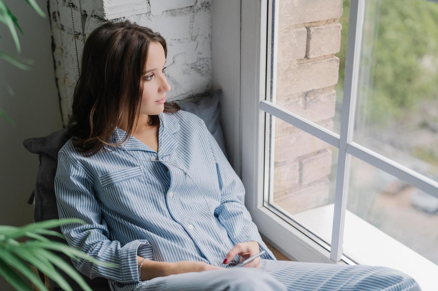 Horizontal shot of thoughtful young woman sits on windowsill, dressed in casual nightwear, uses smart phone and earphones, listens calm music while looks thoughtfully in window, enjoys home time photo