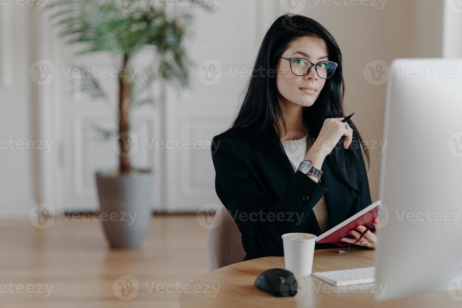 Photo of serious woman writes information in notebook, studies new online business course, poses in coworking space with computer, works on project, writes some ideas. Business and work concept