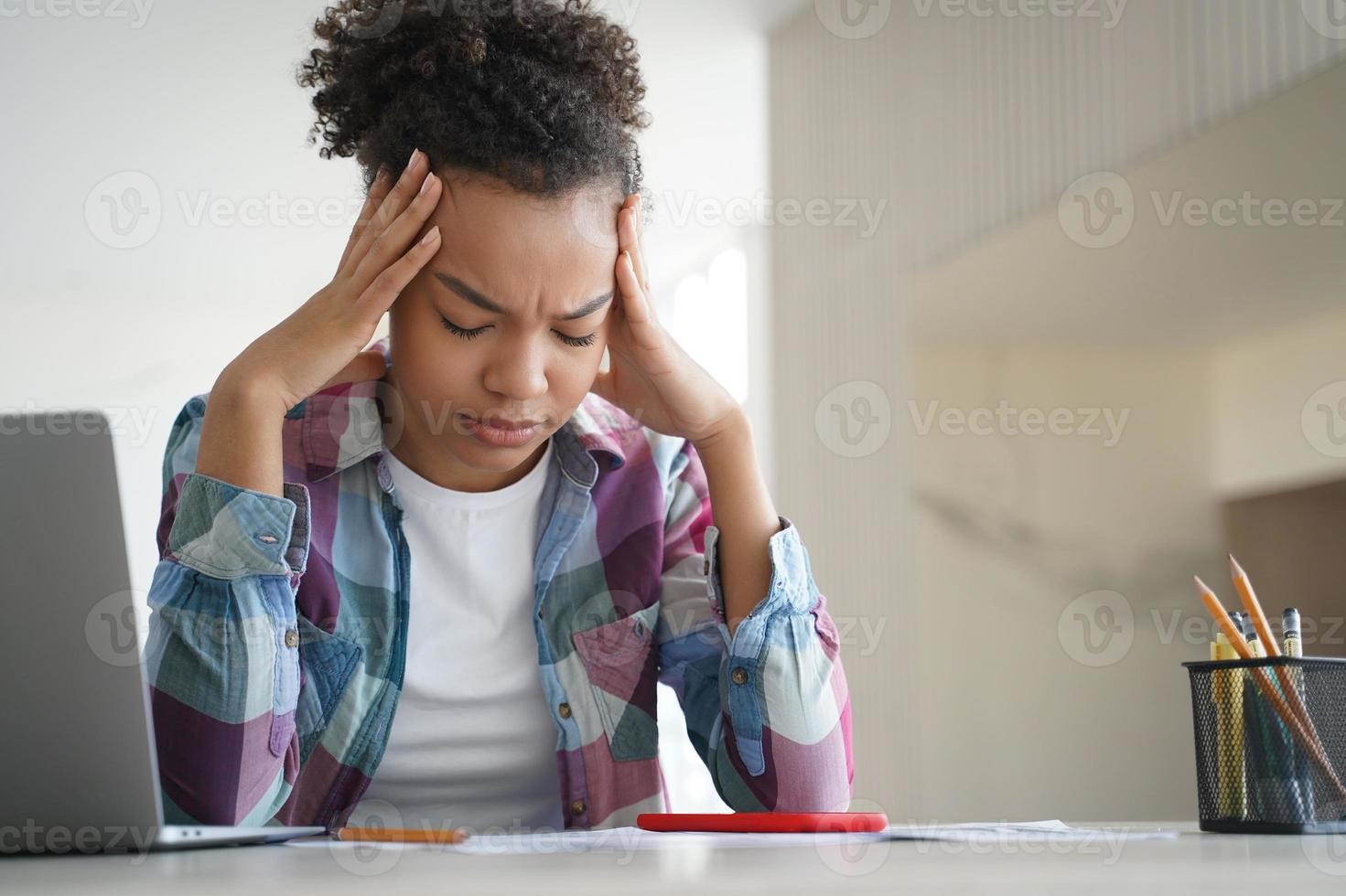 una adolescente afroamericana cansada sufre dolor de cabeza haciendo los deberes en una laptop. aprendizaje electrónico difícil foto