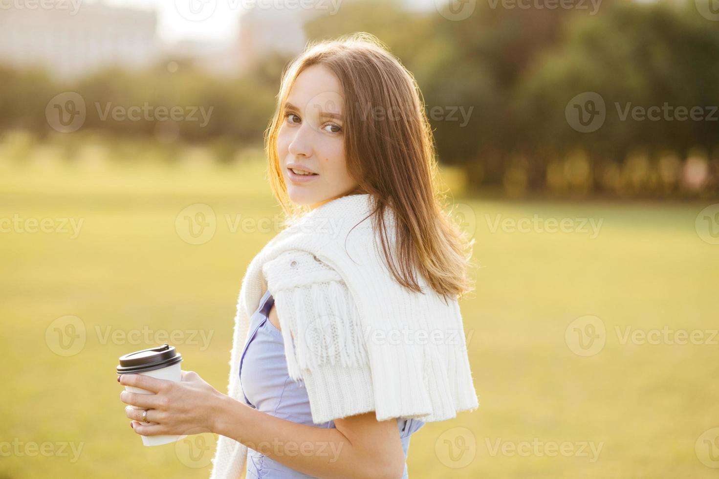 Sideways shot of pretty young girl with dark har, holds cup with coffee or cappuccino, breathes fresh air, enjoys calm atmosphere, admires beautiful nature and sunset. People and lifestyle concept photo