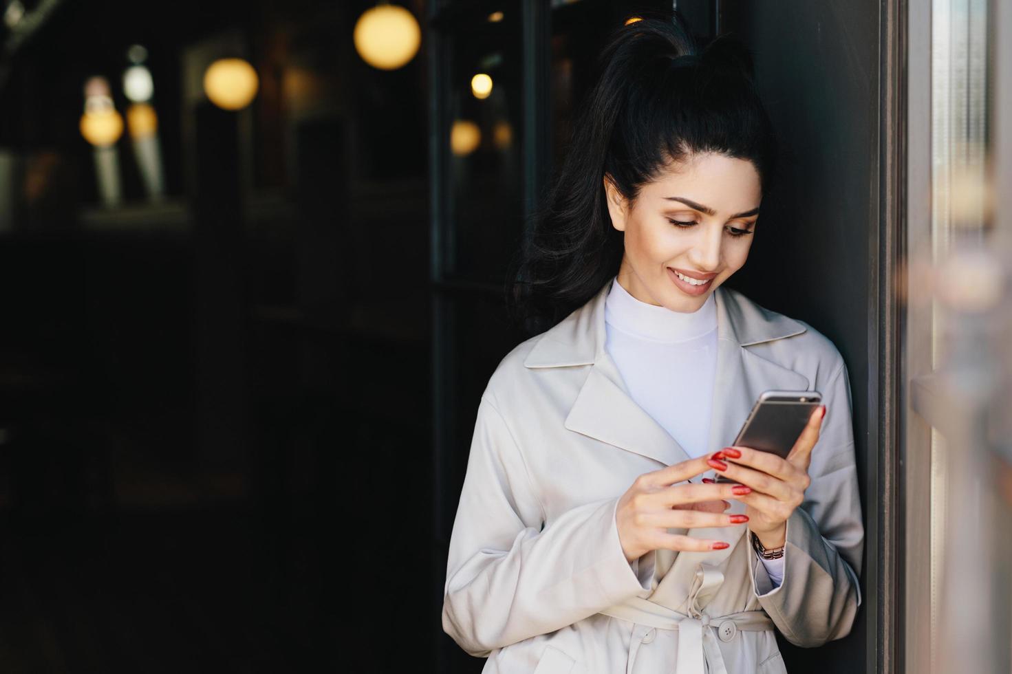 Fashionable beautiful woman with dark hair tied in pony tail dressed in white elegant coat holding cell phone looking into screen with delightful expression. Young pretty model communicating online photo