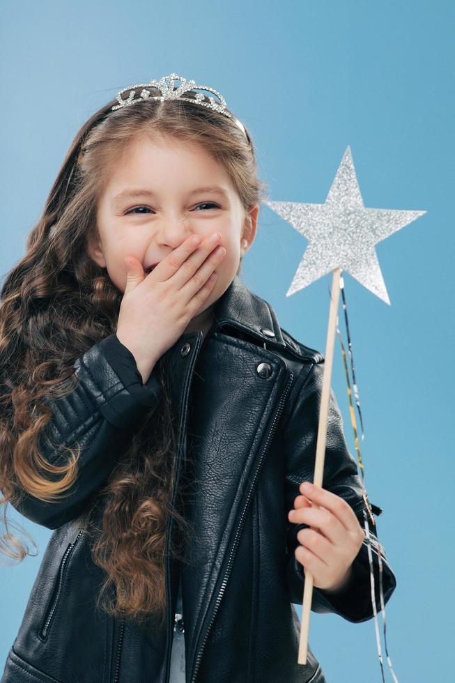 Overemotive positive small child covers mouth with one palm, wears fashionable black leather jacket and crown, holds magic wand in hand, isolated over blue background. Female kid poses indoor photo