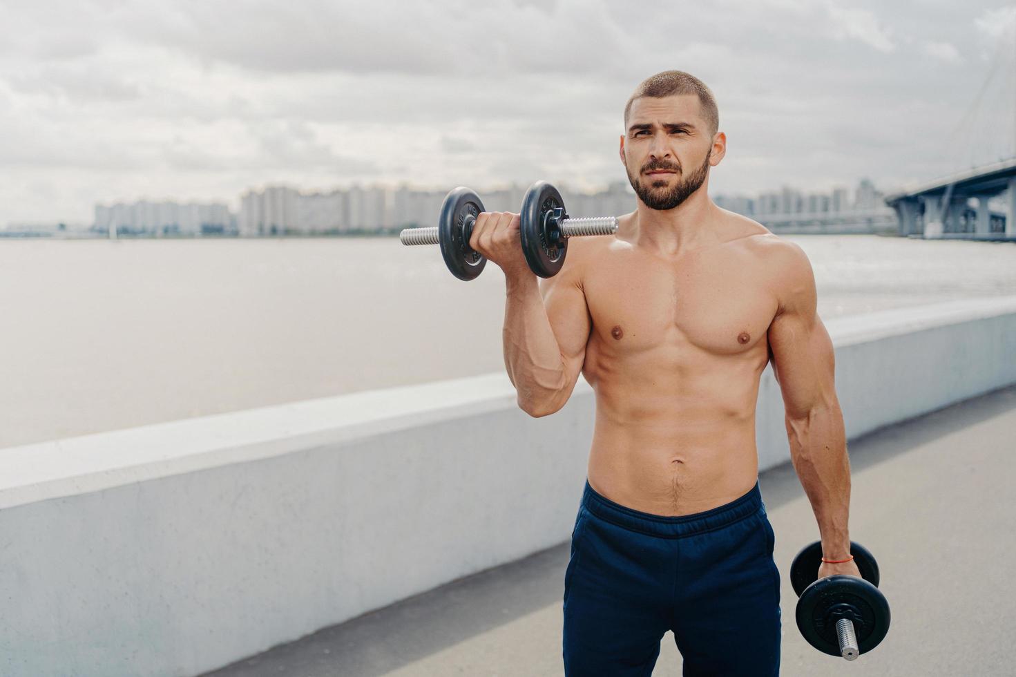 Handsome bearded man pumps up muscles, does exercises with barbells, leads sporty lifestyle, has muscular strong body, stands outdoor near river. Bare chested bodybuilder lifts heavy dumbbells photo
