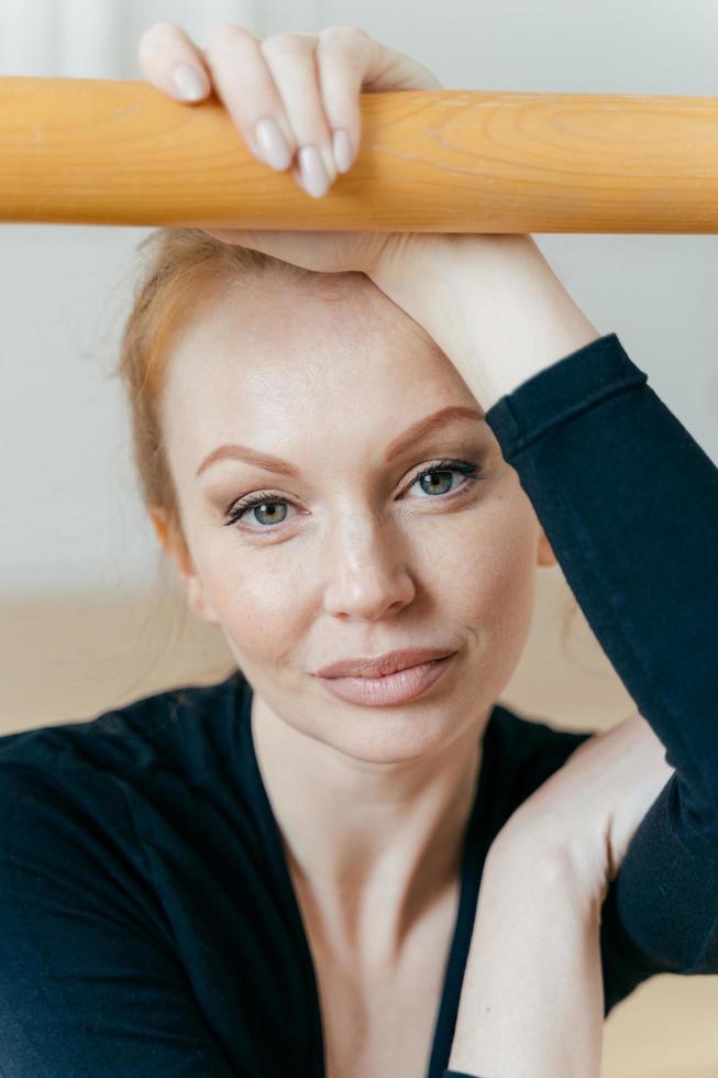 Close up shot of restful woman with ginger hair, has make up, looks at camera, has serious expression, holds hand on handrail, busy doing gymnastic exercises, has natural beauty. Ballet concept photo
