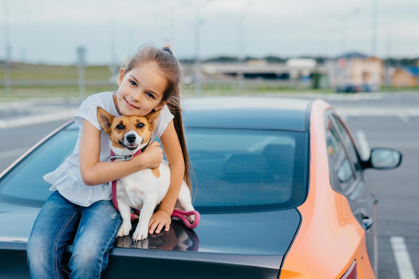 Small attractive female child embraces her favourite dog, sit together at trunk of car, have rest after stroll, enjoy summer day, have friendly relationship. Children, pets and lifestyle concept. photo