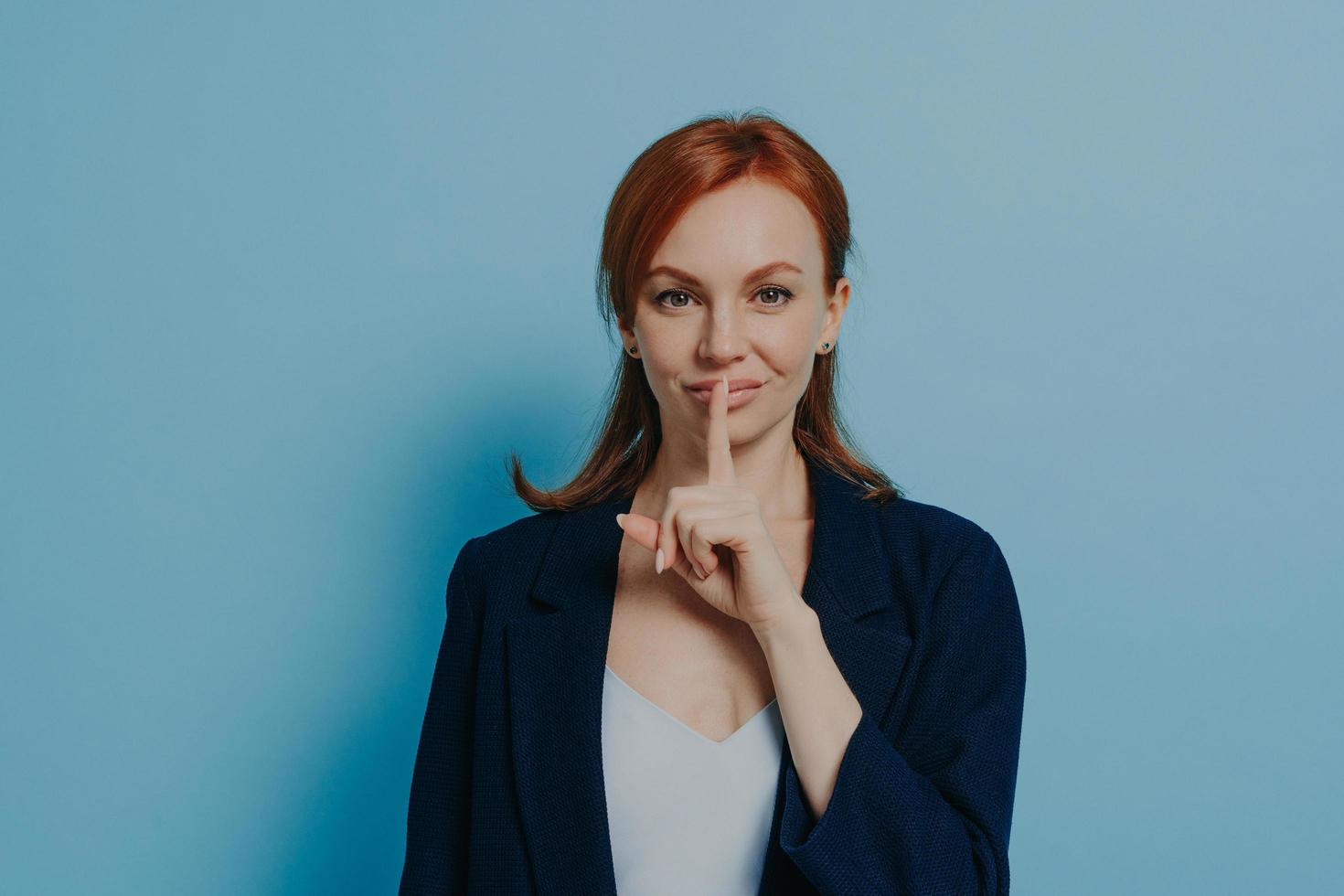 Young redhaired woman asking to keep silence or not to tell secret, isolated on blue background photo