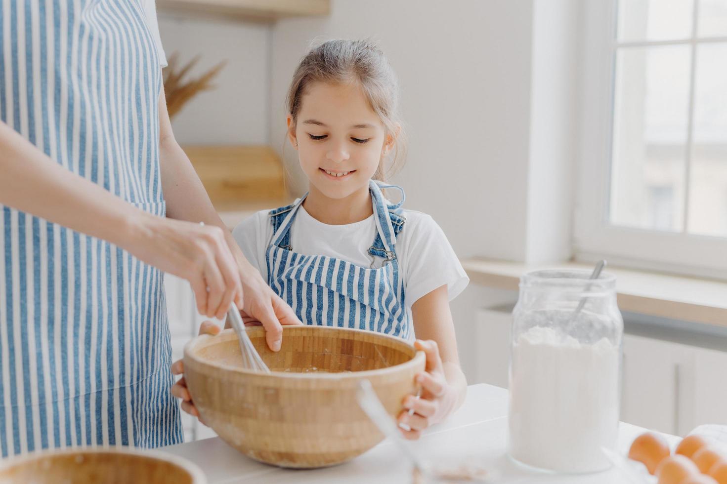 Curious small female helper looks attentively how mother cooks, helps to whisk ingredients, wears white t shirt and striped apron, prepare tasty cake together, like baking something delicious photo