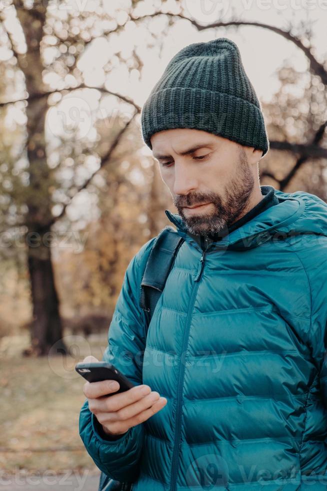 Vertical shot of concentrated hipster guy with thick beard, wears anorak and hat, holds cellular, uses special application for finding route, message with friend, breathes fresh air. Lifestyle concept photo