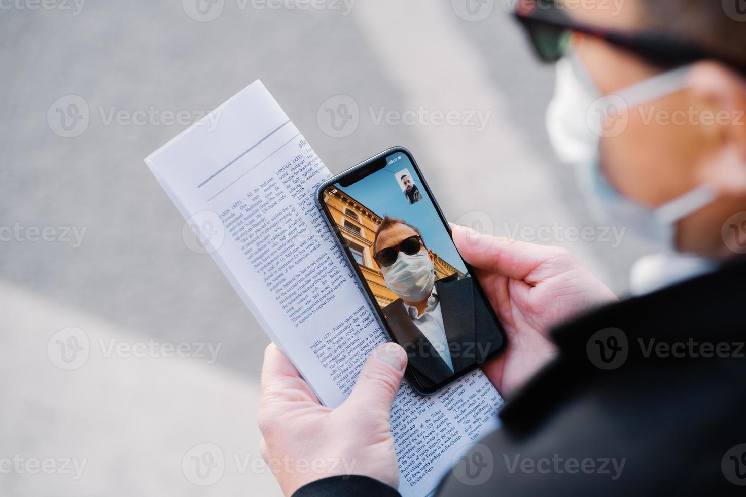 Cropped image of faceless man makes video call via modern cellphone, wears protective medical mask during virus spreading, has only distance communication to prevent coronavirus, holds newspaper photo