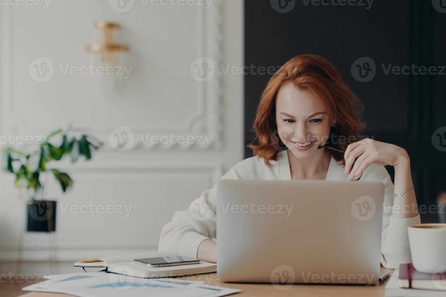 Positive happy female freelancer has busy working day, works distantly from home, sits in front of laptop computer against modern interior, works on creative task, watches webinar for improving skills photo